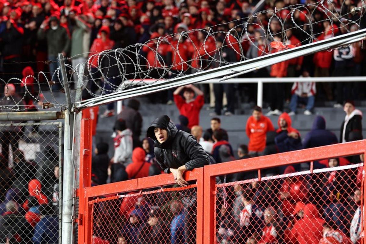 A fan of Independiente climbs a gate during the interruption of the Copa Sudamericana round of 16 second leg football match between Argentina's Independiente and Chile's Universidad de Chile at the Libertadores de America stadium in Avellaneda, Buenos Aires province, Argentina on August 20, 2025. The match between Independiente and Universidad de Chile was suspended due to incidents in the stands during the second half, according to the Argentine club and confirmed by Conmebol. The score was tied 1-1. Alejandro PAGNI / AFP